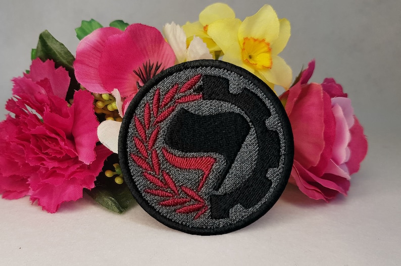 May include: A black and gray embroidered patch with a red stylized fist and a wreath around it. The patch is circular and has a gear design on the right side.