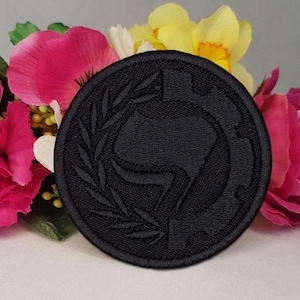 Puede incluir: Un parche redondo bordado en negro con un diseño central dentro de una corona de laurel. El parche se encuentra sobre un fondo de vibrantes flores rosas, amarillas y blancas, creando un contraste llamativo.