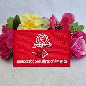 May include: A rectangular red embroidered patch featuring a white stitched design of a rose above two hands shaking. The text "Democratic Socialists of America" is embroidered in white at the bottom of the patch, which is centered in front of flowers.