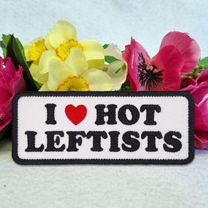 May include: A rectangular embroidered patch with a white background and a black border. The text reads I (with a red heart symbol) HOT LEFTISTS in bold black capital letters. The patch is positioned in front of a cluster of yellow and pink flowers.