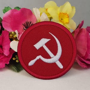 以下が含まれることがあります： 白い鎌とハンマーのエンブレムが付いた、丸い赤いパッチ。 パッチは赤い縁で囲まれ、ピンク、黄色、白の花を背景に配置されています。 これは刺繍されたアイテムである可能性があります。