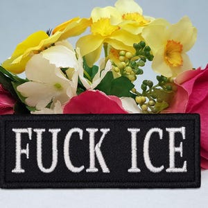 Pode incluir: Um patch bordado preto com as palavras "FUCK ICE" em letras brancas. O patch é colocado contra um fundo de flores coloridas, incluindo flores rosa, amarelas e brancas.