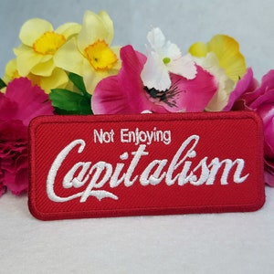 Op de afbeelding: Rode geborduurde patch met witte tekst die "Not Enjoying Capitalism" luidt. De patch is omringd door roze en gele bloemen.