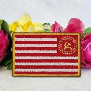 May include: Embroidered patch featuring a red and white striped design with a gold border. The upper right corner displays a gold hammer and sickle emblem within a circular design. The patch is rectangular and is set against a backdrop of pink and yellow flowers.