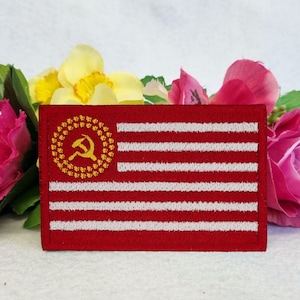 May include: A red and white embroidered patch with a flag-like design. The upper left corner has a gold hammer and sickle emblem within a gold wreath. The rectangular patch features red and white stripes.
