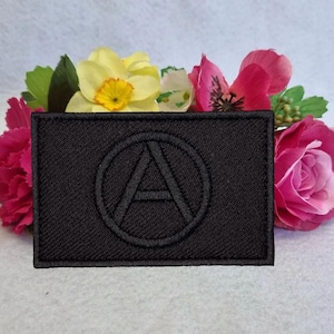 Puede incluir: Un parche rectangular negro con un círculo cosido y la letra "A" en su interior. El parche está rodeado de flores rosas, amarillas y verdes. Probablemente un emblema o un elemento decorativo.