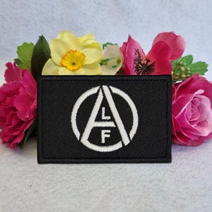 May include: A black patch with a white circular design containing the letters "A", "L", and "F". The patch is surrounded by pink, yellow, and green flowers. The patch is rectangular in shape.