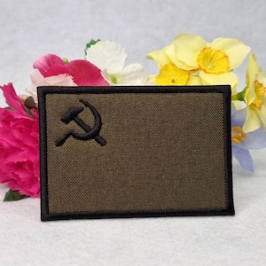 May include: A green embroidered patch with a black hammer and sickle design. The patch has a black border and is surrounded by flowers.