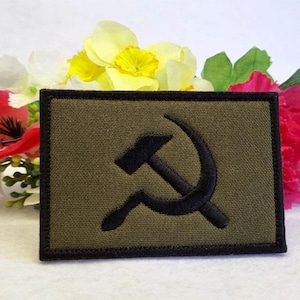 以下が含まれることがあります： 黒い刺繍のハンマーと鎌のシンボルが施された緑色の布のパッチ。パッチは黒い縁取りがされています。