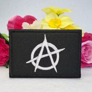 May include: A rectangular black patch featuring a white embroidered anarchy symbol in the center. The symbol consists of a capital letter A enclosed within a circle. The patch has a stitched border and a textured fabric surface.