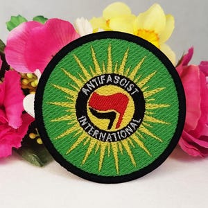 Könnte beinhalten: Ein gestickter Aufnäher in Grün und Gelb mit schwarzem Rand. Der Aufnäher zeigt eine stilisierte Faust mit einem roten Arm und den Text "Anti-Faschistische Internationale".
