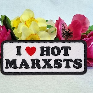 May include: A rectangular embroidered patch with a white background and a black border. The text reads I (with a red heart symbol) HOT MARXSTS in bold black lettering. The patch is positioned in front of a cluster of yellow and pink flowers.