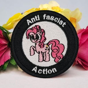 Könnte beinhalten: Gestickter Aufnäher mit schwarzem Rand und den Worten "Anti fascist Action". Der Aufnäher zeigt ein rosa Cartoon-Pony in einem weißen Kreis. Der Aufnäher ist von rosa, gelben und grünen Blumen umgeben.