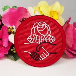 May include: A circular red embroidered patch featuring a white stitched design of a rose positioned above two hands shaking. The embroidery is detailed with clean lines, showcasing a stylized floral motif and a gesture of unity on a solid red background.
