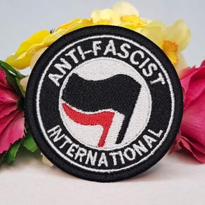 Pode incluir: Um emblema bordado circular com um design de bandeira preta e vermelha centrado num fundo branco. A borda preta contém o texto bordado em branco ANTI-FASCIST INTERNATIONAL. O emblema é exibido contra um fundo de flores.