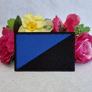 Può includere: Una toppa ricamata blu e nera con una linea diagonale che divide i colori. La toppa è circondata da fiori rosa e gialli.