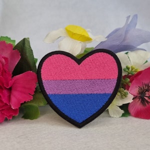 Op de afbeelding: Een hartvormige geborduurde patch met horizontale strepen in roze, paars en blauw. De patch heeft een dikke zwarte rand en is omringd door kunstbloemen in tinten roze, wit, geel en lichtpaars.