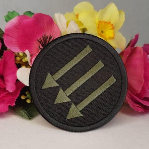 Op de afbeelding: Een ronde geborduurde patch met een zwarte achtergrond en drie olijfgroene pijlen die in een diagonale opstelling naar beneden wijzen. De patch heeft een dikke zwarte gestikte rand en wordt getoond tegen een achtergrond van kleurrijke bloemen.