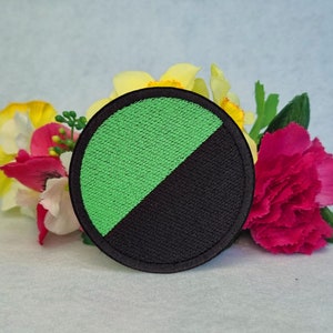 May include: A round patch with a black border, divided diagonally into green and black sections. The patch is embroidered with a textured appearance. The background features colorful flowers.