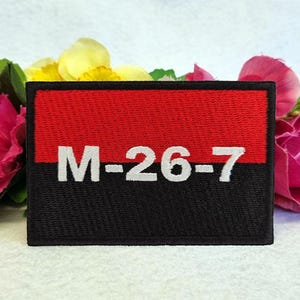 May include: A rectangular embroidered patch featuring a horizontal split design with a red upper half and a black lower half. The white text M-26-7 is stitched across the centre, spanning both the red and black sections of the fabric patch.