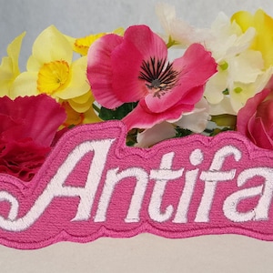May include: A pink embroidered patch with the word "Antifa" in white script, set against a backdrop of colourful artificial flowers. The flowers include pink, yellow, and white blooms.