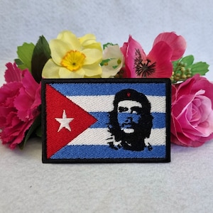 May include: Embroidered patch featuring the Cuban flag with a portrait of Che Guevara. The flag is red, white, and blue with a white star. The patch is surrounded by pink and yellow flowers.