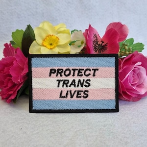 May include: A blue, pink and white embroidered patch with the text "PROTECT TRANS LIVES" on a black background.