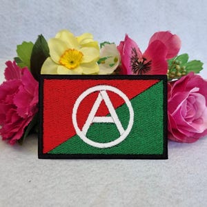 May include: Embroidered patch with a red and green diagonal design, featuring a white "A" within a circle. The patch is set against a backdrop of pink, yellow, and green flowers. The patch has a rectangular shape with a black border.