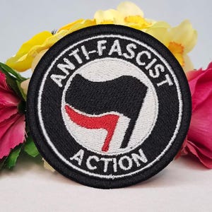 Könnte beinhalten: Gestickter Aufnäher mit den Worten "ANTI-FASCIST ACTION" in Weiß auf schwarzem, kreisförmigem Hintergrund. Der Aufnäher zeigt eine schwarze Flagge mit einem roten Element. Der Aufnäher ist von rosa, gelben und grünen Blumen umgeben.