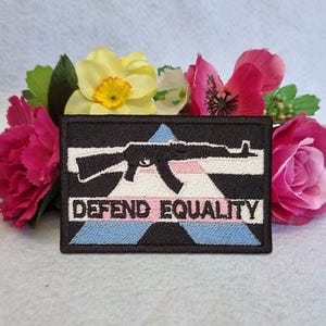 Pode incluir: Um emblema retangular bordado com borda preta apresentando a silhueta de um rifle preto centralizada sobre um triângulo de listras azul-claro, rosa e branco. O texto DEFEND EQUALITY está bordado em letras maiúsculas pretas abaixo.