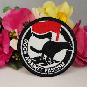 May include: Black and white embroidered patch with a red silhouette of a fist. The patch says "Dogs Against Fascism" in a circle.