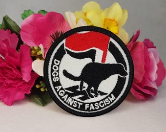May include: Black and white embroidered patch with a red silhouette of a fist. The patch says "Dogs Against Fascism" in a circle.