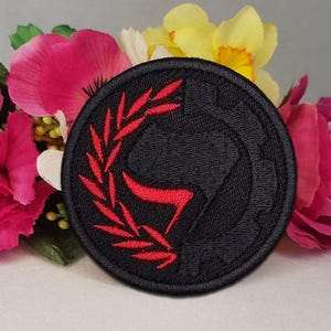 Könnte beinhalten: Ein schwarzer runder Aufnäher mit einem roten Emblem, das eine stilisierte Figur und einen Lorbeerkranz zeigt. Der Aufnäher ist von rosa, gelben und weißen Blumen umgeben, was einen lebendigen Kontrast erzeugt.
