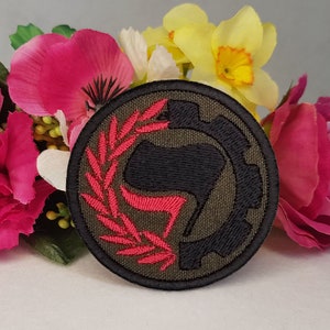 以下が含まれることがあります： スタイリッシュな拳と花輪が刺繍された、丸い緑、黒、赤の刺繍パッチ。パッチはピンクと黄色の花に囲まれています。