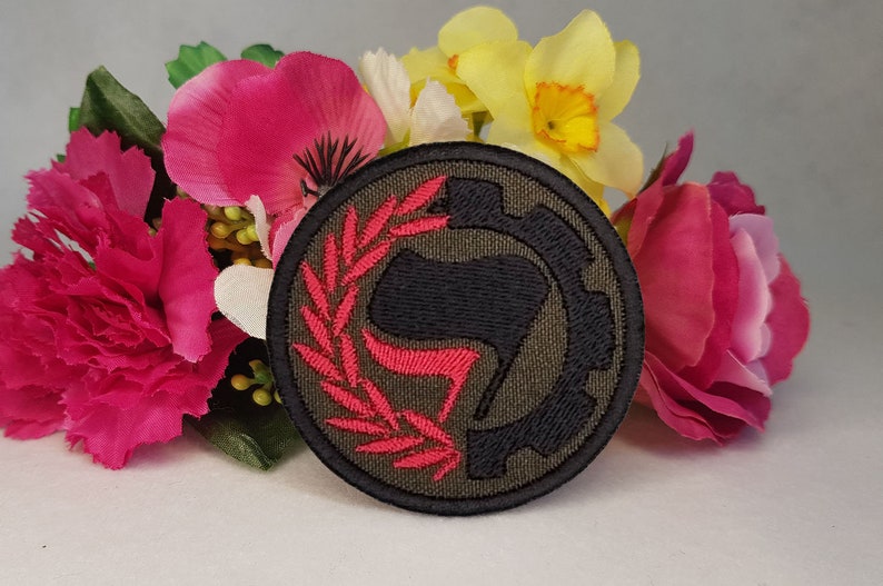May include: A round, green, black, and red embroidered patch with a stylized fist and a wreath. The patch is surrounded by pink and yellow flowers.