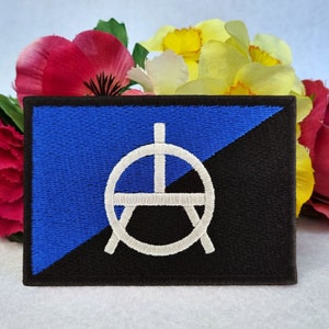 May include: A blue and black embroidered patch featuring a white anarchy symbol within a circle. The patch is surrounded by pink and yellow flowers.