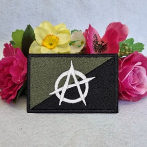 May include: A black and green embroidered patch with a white anarchy symbol. The patch is surrounded by pink and yellow flowers.