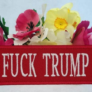 Op de afbeelding: Een rechthoekige rode geborduurde patch met de witte tekst FUCK TRUMP in een vetgedrukt schreeflettertype. De patch is geplaatst voor een boeket kunstbloemen, waaronder roze anjers, een gele narcis en witte bloesems.