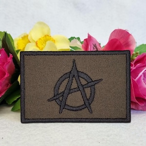 Op de afbeelding: Een rechthoekige geborduurde patch met een donker olijfgroene achtergrond en een zwarte rand. In het midden van de patch staat een zwart geborduurd anarchiesymbool, bestaande uit een hoofdletter A in een cirkel.