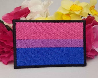 May include: A rectangular embroidered patch featuring three horizontal stripes in pink, purple, and blue, enclosed by a thick black border. The design displays the bisexual pride flag colors with a textured, stitched surface and clean edges.