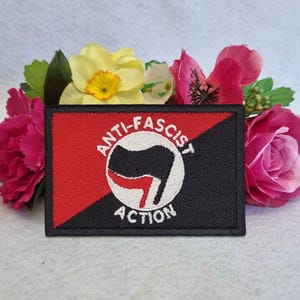 May include: A rectangular embroidered patch featuring a diagonal split of red and black. In the center, a white circle contains a black and red flag icon with the text ANTI-FASCIST ACTION arched around the emblem in white capital letters.