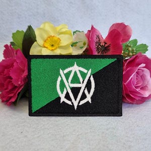 以下が含まれることがあります： 緑と黒の刺繍パッチに白い無政府主義のシンボルが描かれています。パッチはピンクと黄色の花に囲まれています