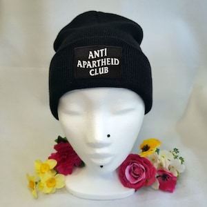 May include: Black beanie with a white patch that reads "ANTI APARTHEID CLUB".