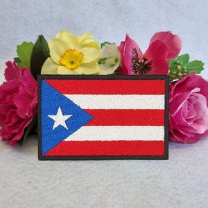 May include: An embroidered patch featuring the flag of Puerto Rico. The flag is red, white, and blue with a white star in the blue triangle. The patch is surrounded by a black border.