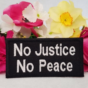 Könnte beinhalten: Schwarzer gestickter Aufnäher mit weißem Text, der "No Justice No Peace" lautet. Der Aufnäher ist von rosa und gelben Blumen umgeben.