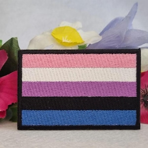 May include: A transgender pride flag patch with five horizontal stripes in pink, white, light blue, purple, and black. The patch is embroidered and has a black border.