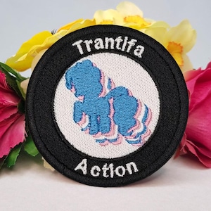 Könnte beinhalten: Gestickter Aufnäher mit schwarzem Rand, weißer Mitte und blau-rosa Grafik. Der Aufnäher trägt den Text "Trantifa" über der Grafik und "Action" darunter. Der Aufnäher ist von rosa, gelben und grünen Blumen umgeben.