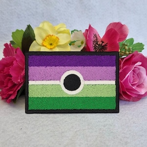 May include: Embroidered patch with the non-binary pride flag design. The flag has horizontal stripes in purple, light purple, white, green, and a black circle in the centre. The patch is framed by a black border, surrounded by pink and yellow flowers.