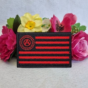 Puede incluir: Un parche bordado rojo y negro con un símbolo de anarquía en un círculo. El parche tiene la forma de una bandera estadounidense.