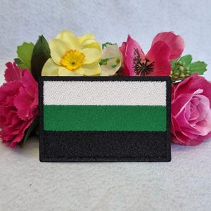 May include: A rectangular patch with a three-stripe design in white, green, and black. The patch is surrounded by pink and yellow flowers with green leaves. The patch is likely an emblem or flag.
